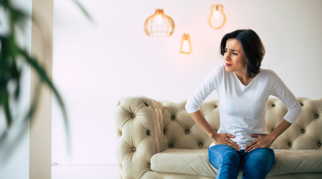A woman sits on a couch, clutching her stomach in pain. Learn why immune booster vitamins are important for your health